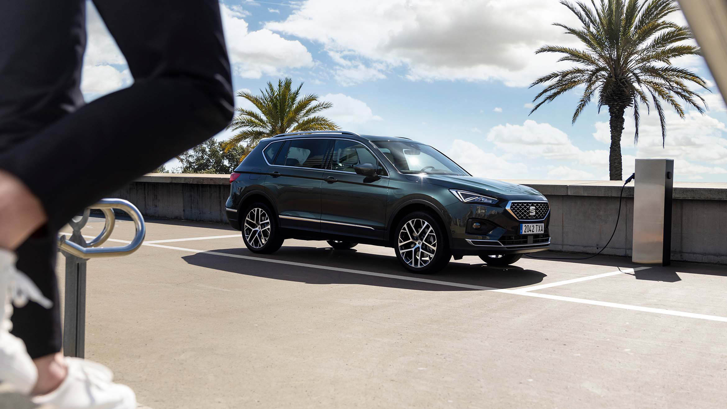 Inimene kõnnib laadima pandud uue SEAT Tarraco XPERIENCE poole, kerevärv Dark Camouflage  