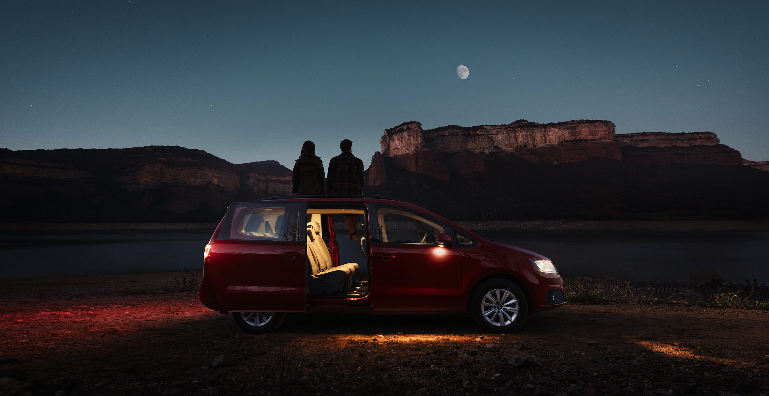 Mahtuniversaal SEAT Alhambra lahtiste ustega külgvaates õhtupimeduses ning kaks inimest vaatamas kuud ja mägist maastikku